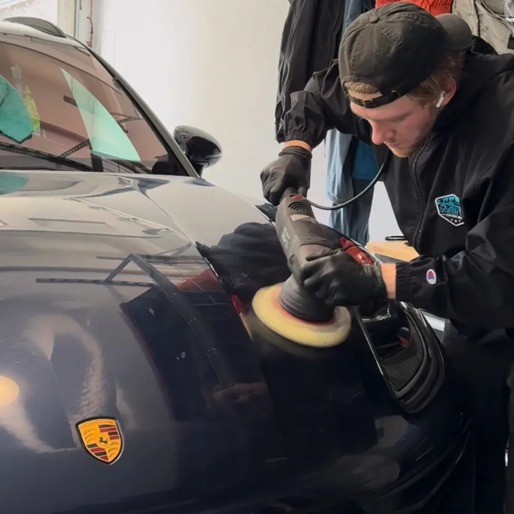 Machine polisher with a foam pad working the front end of a dark blue Porsche
